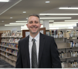 man with beard wearing suit smiling in front of book stacks