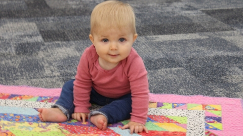 infant child looking at teh camera with blonde hair wearing a pink long sleeve shirt and dark pants with bare feet sitting on a quilt 