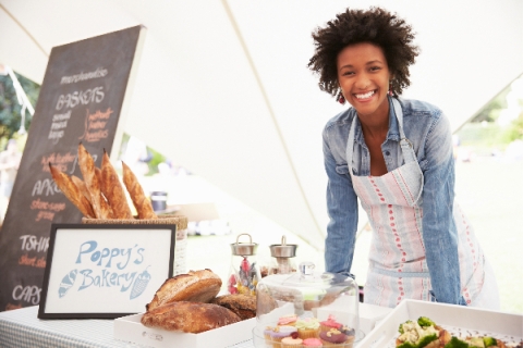 african american woman smiling, wearing a white apron, leaning on a table of baked goods at a farmers market