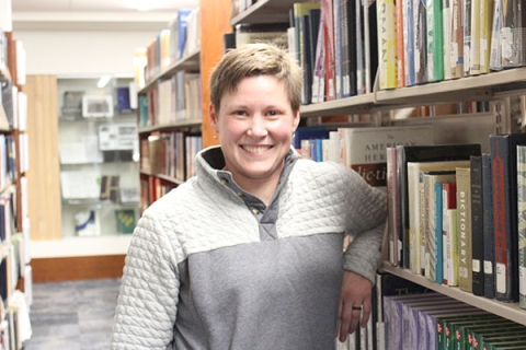 woman with short blond hair wearing a gray two-toned sweatshirt leaning on a bookshelf