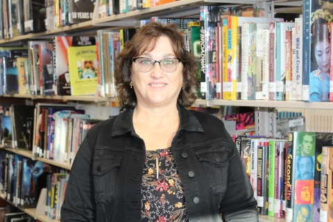 woman with dark hair and glasses wearing a black denim jacket leaning against a bookshelf