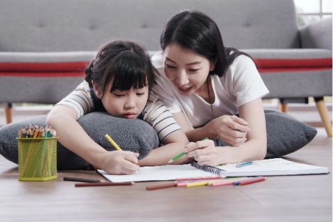 asian mom laying on pillow with small asian child working on homework