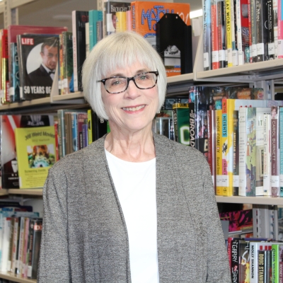 woman with gray hair and glasses wearing a gray sweater cardigan standing next to a bookshelf