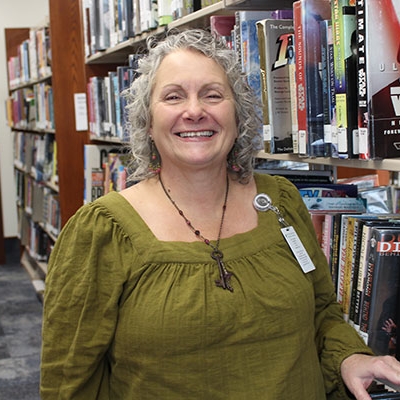 woman with gray curly hair wearing a green blouse standing next to a book shelf